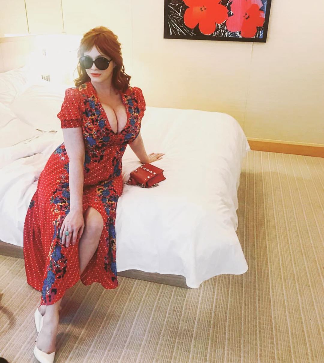 Mommy Christina in a red floral dress and large sunglasses, sitting on a hotel room bed.
